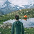Mann mit einem grünen Kapuzenpullover von hinten, der auf ein Bergpanorama blickt, vor ihm ein See und ein oranges Zelt